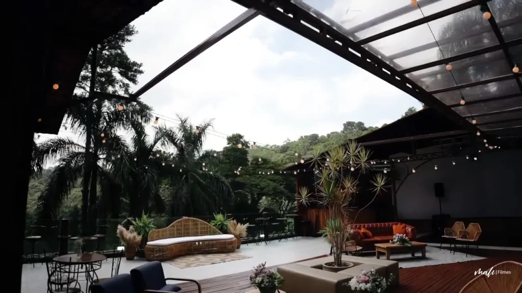 Amplo espaço rústico-chique para casamento em Mairiporã, apresentando um lounge com sofás elegantes, teto translúcido com cordões de luzes e vista panorâmica para a natureza.