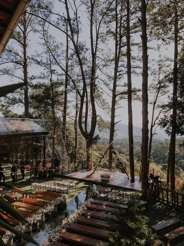 Cenário ao ar livre para casamento em Mairiporã, com altar sobre um deck de madeira rodeado por pinheiros altos, caminho espelhado refletindo a luz, bancos rústicos ladeados por flores brancas e vista panorâmica para as montanhas.
