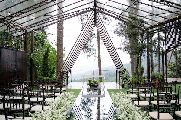 Altar de casamento rústico sob uma estrutura de vidro no Villa Vezzane. O chão espelhado reflete os arranjos de flores brancas que ladeiam o caminho. Ao fundo, o altar com piso de madeira, mesa para celebrante e vista para o horizonte. Cadeiras escuras estão dispostas para os convidados em um gramado verde.