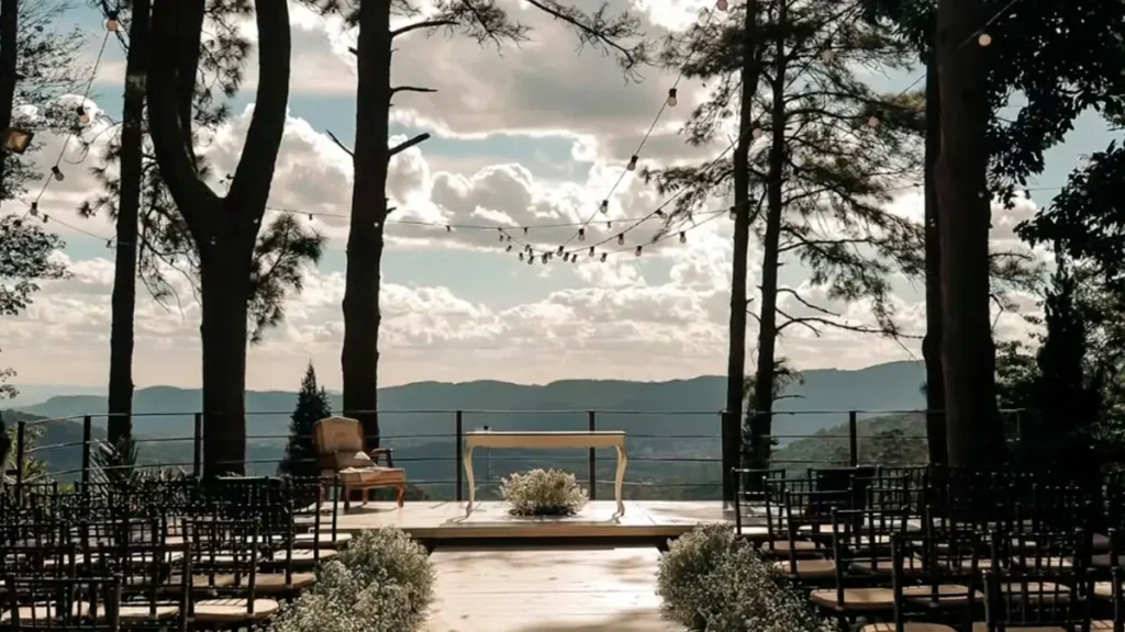 Altar de casamento rústico ao ar livre no Villa Vezzane, com deck de madeira, mesa para celebrante, cadeiras para os convidados e uma vista deslumbrante para a paisagem de Mairiporã ao fundo, sob um céu com nuvens. Árvores altas emolduram a cena e um cordão de luzes está suspenso acima do altar