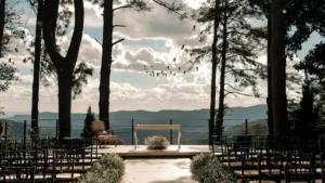 Altar de casamento rústico ao ar livre no Villa Vezzane, com deck de madeira, mesa para celebrante, cadeiras para os convidados e uma vista deslumbrante para a paisagem de Mairiporã ao fundo, sob um céu com nuvens. Árvores altas emolduram a cena e um cordão de luzes está suspenso acima do altar