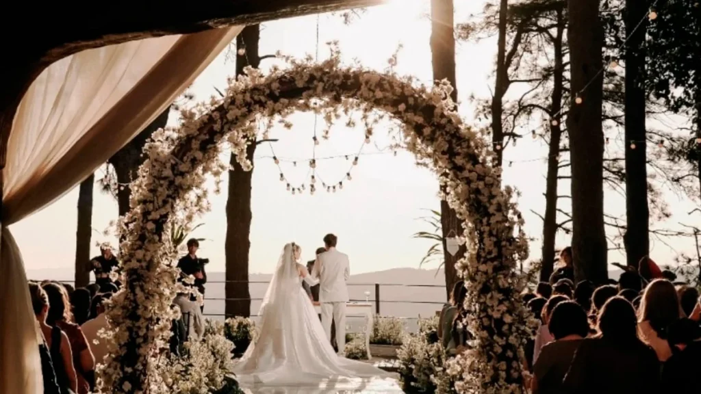 Cerimônia ao ar livre no dia do casamento, onde os noivos estão em pé no altar, vistos de costas e emoldurados por um grande arco de flores brancas. A noiva veste um vestido branco longo com véu e o noivo usa um terno claro. O cenário rústico é rodeado por árvores altas, iluminado pela luz do sol ao fundo e decorado com luzinhas, enquanto os convidados assistem sentados à celebração.