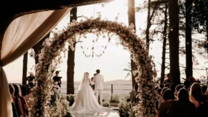 Cerimônia ao ar livre no dia do casamento, onde os noivos estão em pé no altar, vistos de costas e emoldurados por um grande arco de flores brancas. A noiva veste um vestido branco longo com véu e o noivo usa um terno claro. O cenário rústico é rodeado por árvores altas, iluminado pela luz do sol ao fundo e decorado com luzinhas, enquanto os convidados assistem sentados à celebração.